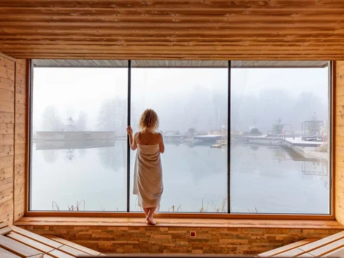 Blog – Neuigkeiten aus dem Pfalzblick Wald Spa Resort Frau in Handtuch blickt aus Holzsauna auf nebligen Teich