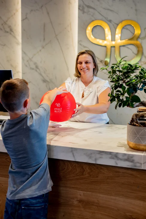 Ihr Hotel im Dahner Felsenland: Pfalzblick Wald Spa Resort Kind bekommt roten Ball an einer modernen Rezeption übergeben