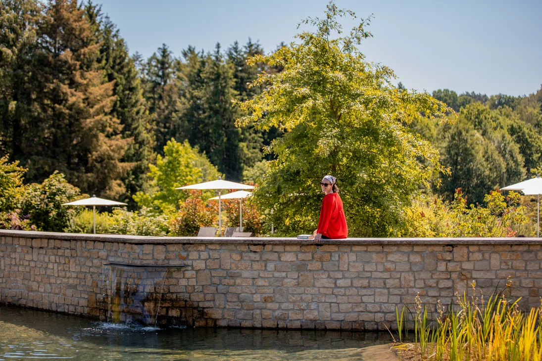 Ihr Hotel im Dahner Felsenland: Pfalzblick Wald Spa Resort Frau in rotem Mantel sitzt an einem Teich mit Grün und Bäumen im Hintergrund