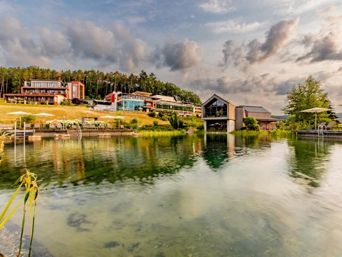Ihr Hotel im Dahner Felsenland: Pfalzblick Wald Spa Resort Moderne Gebäude an einem ruhigen See mit bewölktem Himmel im Hintergrund