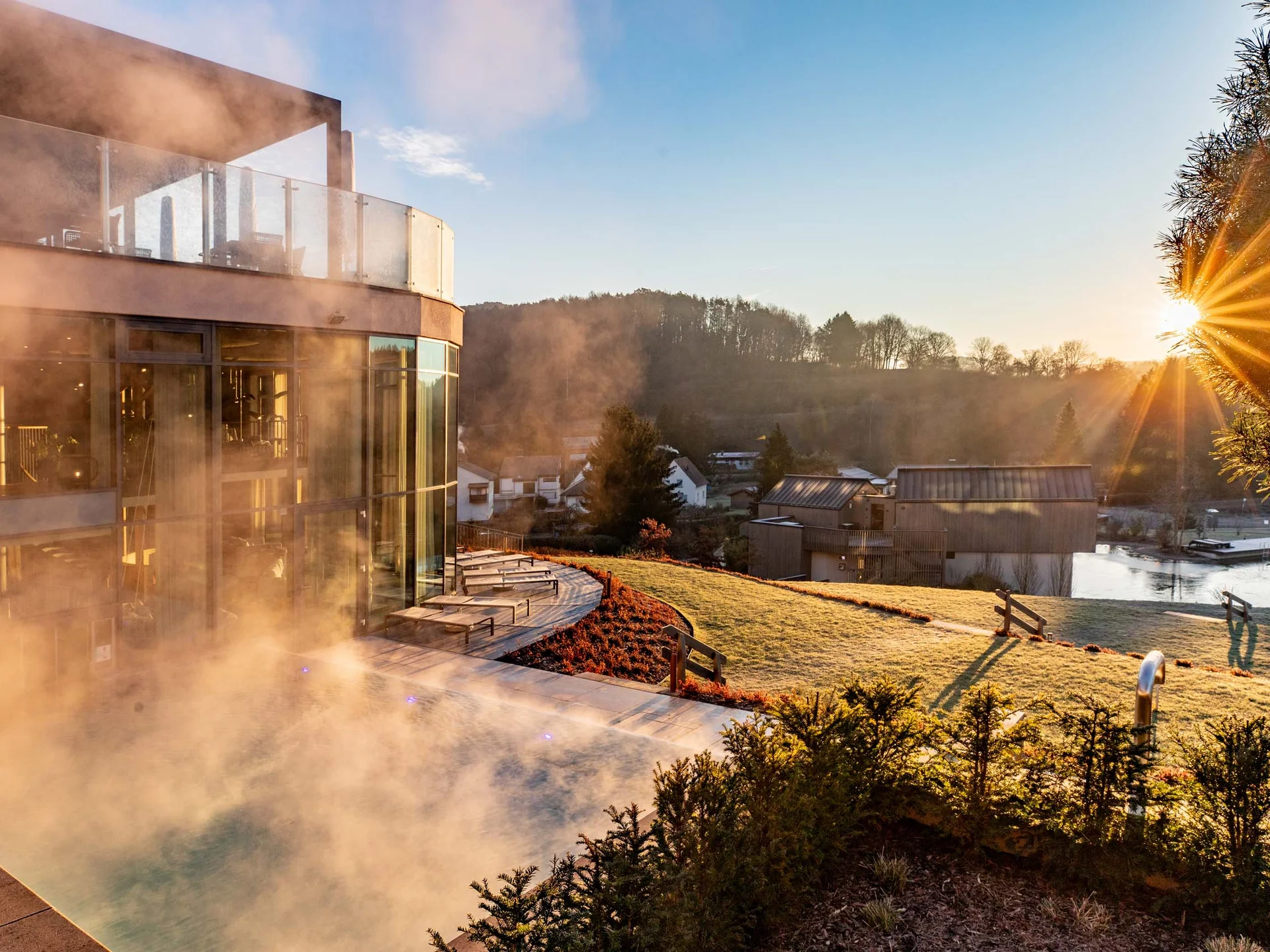 Ihr Hotel im Dahner Felsenland: Pfalzblick Wald Spa Resort Dampf steigt von einem Pool neben einem modernen Gebäude bei Sonnenaufgang im Grünen