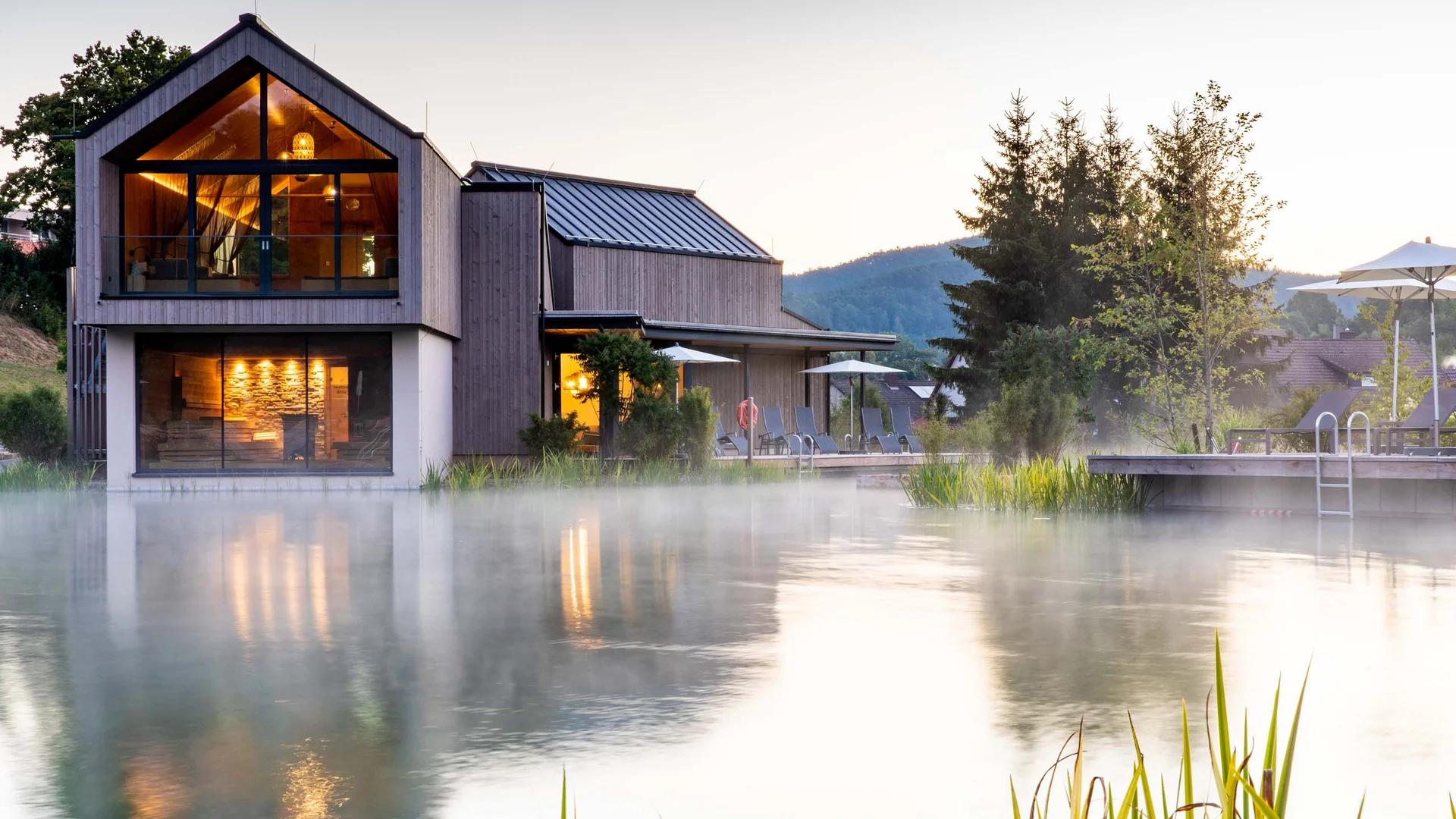 Ihr Hotel im Dahner Felsenland: Pfalzblick Wald Spa Resort Modernes Haus am nebligen See mit beleuchteten Innenräumen und Liegestühlen