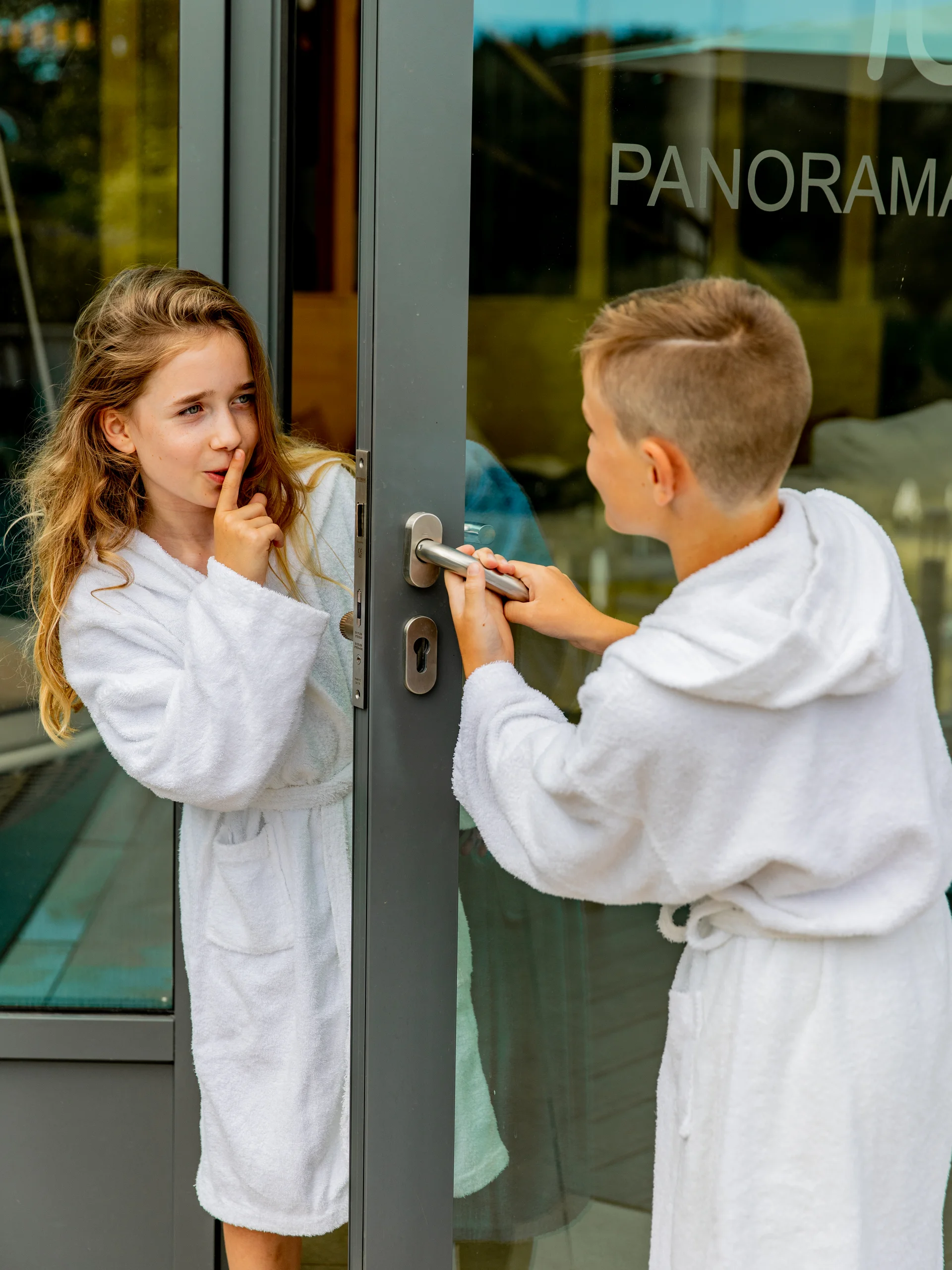 Ihr Hotel im Dahner Felsenland: Pfalzblick Wald Spa Resort Kinder in Bademänteln an einer Glastür, Mädchen macht leise Geste