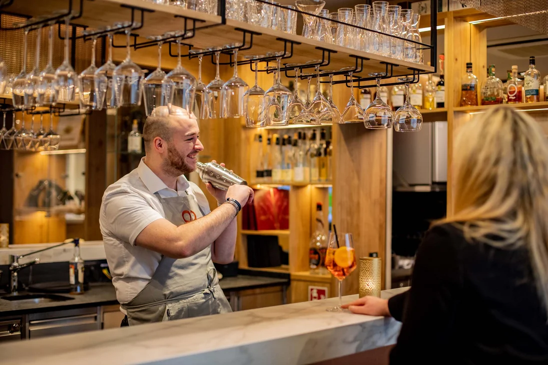Ihr Hotel im Dahner Felsenland: Pfalzblick Wald Spa Resort Barkeeper schüttelt Cocktail hinter der Bar, Frau wartet mit Getränk am Tresen