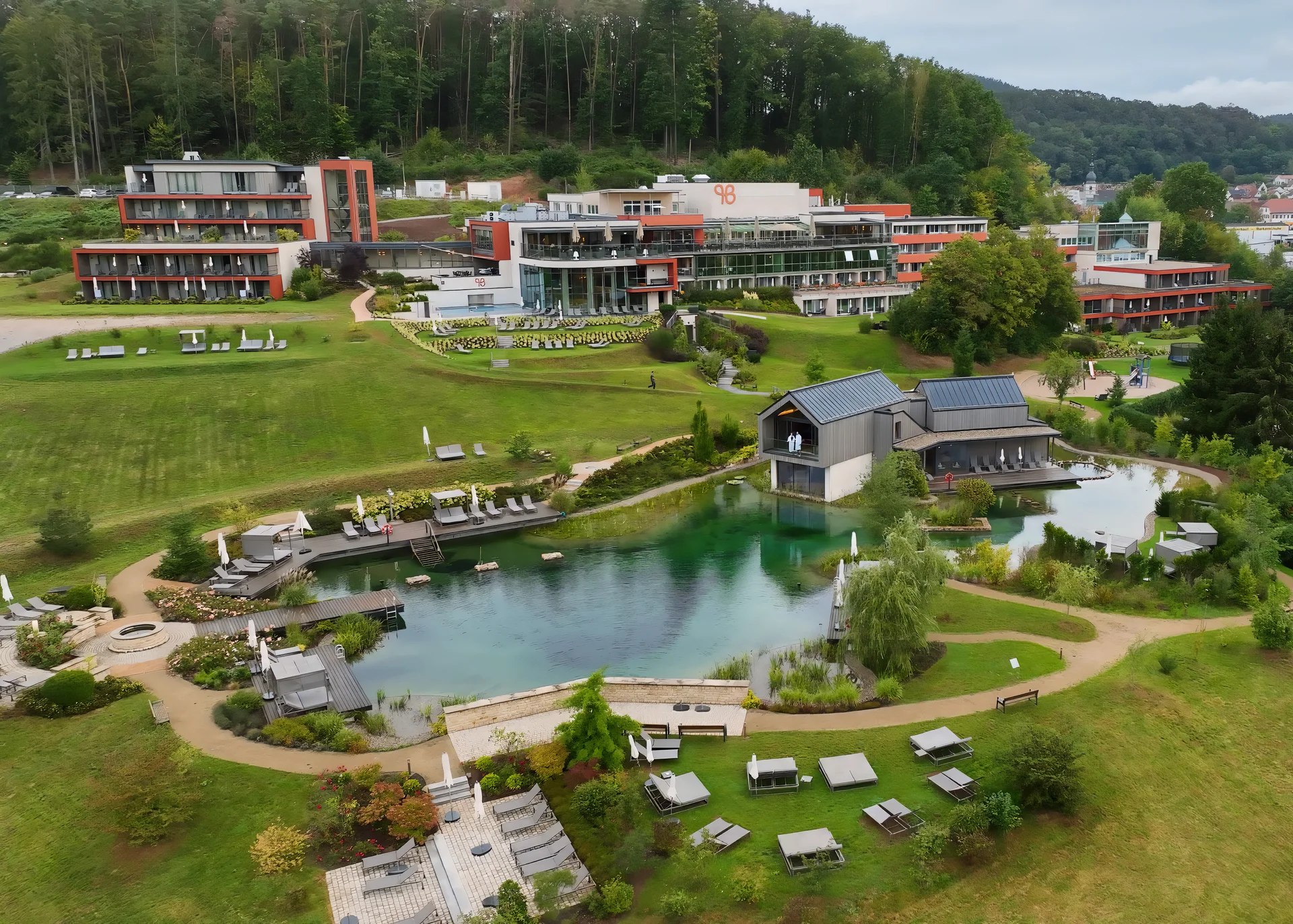 Ihr Hotel im Dahner Felsenland: Pfalzblick Wald Spa Resort Hotelanlage mit Teich, Liegestühlen und umliegender Natur in einer kleinen Stadt