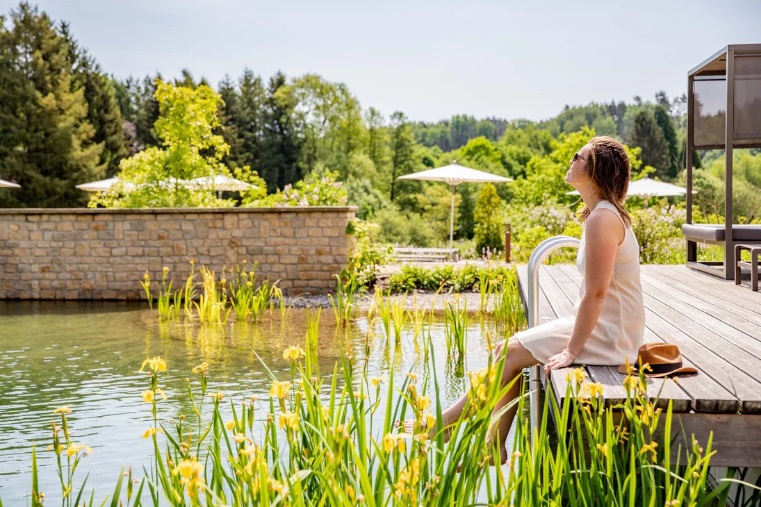 Ihr Hotel im Dahner Felsenland: Pfalzblick Wald Spa Resort Frau entspannt am Holzdeck neben einem natürlichen Schwimmteich mit grünem Umfeld