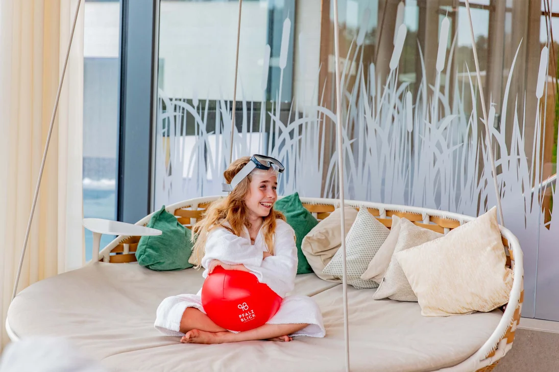 Ihr Hotel im Dahner Felsenland: Pfalzblick Wald Spa Resort Mädchen im Bademantel mit Schwimmbrille sitzt auf Sofa und hält roten Ball