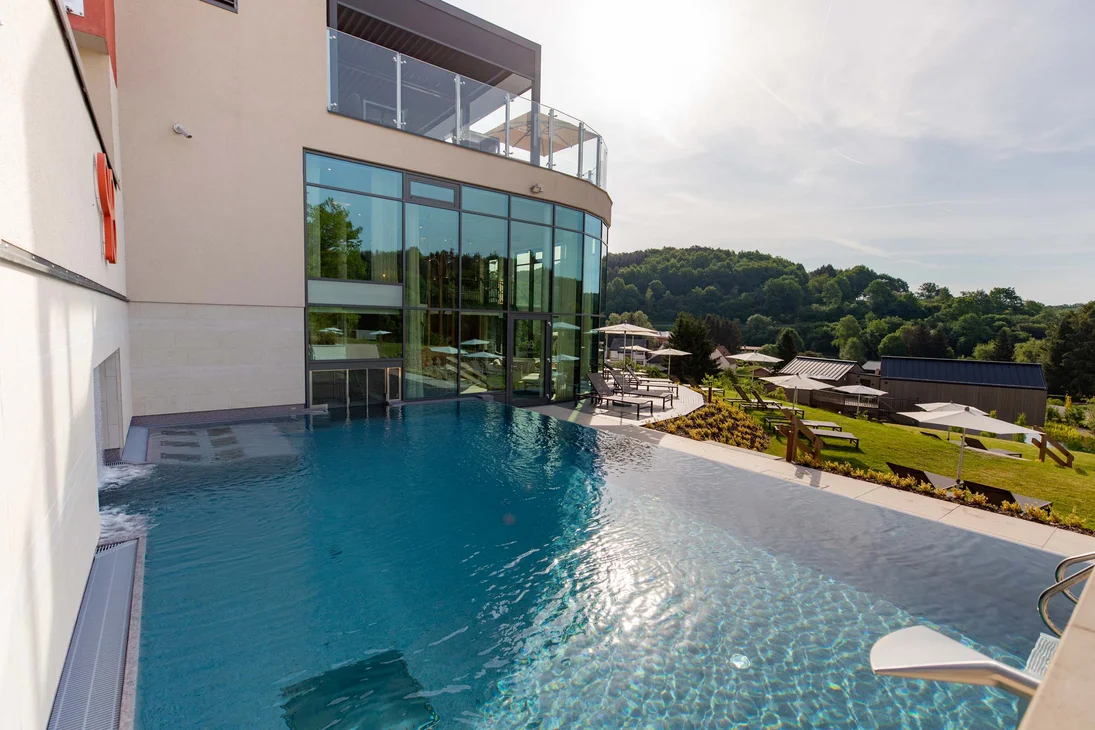 Ihr Hotel im Dahner Felsenland: Pfalzblick Wald Spa Resort Außenpool neben moderne Glasfassade mit Liegestühlen und grüner Landschaft