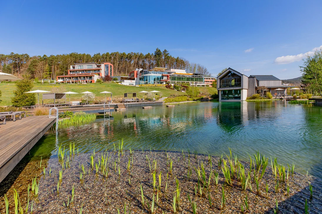 Ihr Hotel im Dahner Felsenland: Pfalzblick Wald Spa Resort Hotelgebäude und Steg am natürlichen Schwimmteich mit grünen Pflanzen unter klarem Himmel